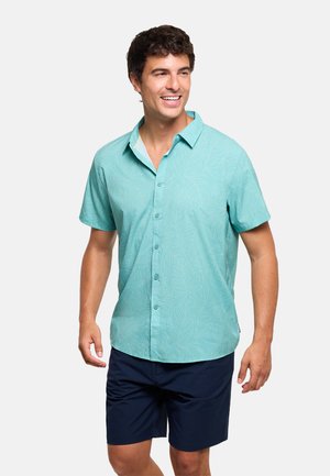 Hombre sonriente con camisa de manga corta azul claro con sutiles patrones de hojas y pantalones cortos azul oscuro sobre un fondo liso.