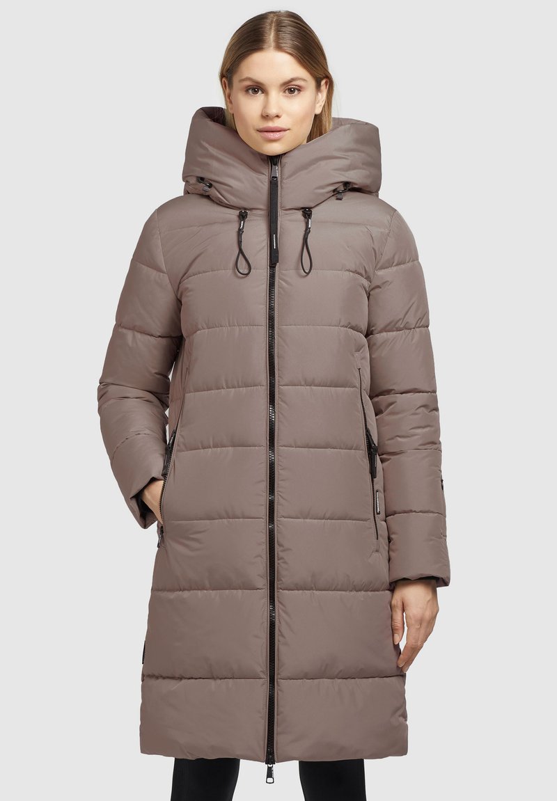Cappotto imbottito di colore marrone tenue, lungo fino al ginocchio, con colletto alto e cappuccio rimovibile. Presenta due tasche laterali e una chiusura a zip integrale.
