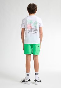 Bílý tričko s barevným grafickým motivem palem a auta na zadní straně, spárované s jasně zelenými šortkami, bílými ponožkami a botami s černými akcenty.