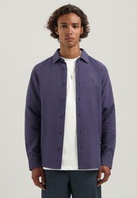 Jonge man met krullend haar draagt een paarse overhemd over een wit t-shirt en donkere broek, staande tegen een lichtgrijze achtergrond.
