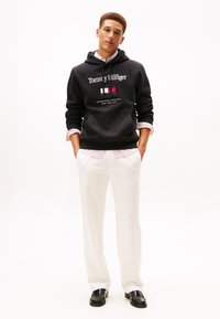 Felpa nera con cappuccio con testo e logo bianchi, abbinata a pantaloni bianchi e mocassini neri. Presenta una camicia a righe sotto.