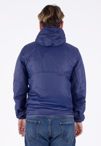 Giacca leggera blu navy con cappuccio, caratterizzata da una finitura liscia e lucida e da polsini elasticizzati, abbinata a jeans in denim blu.