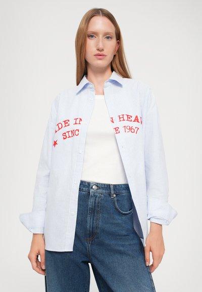 Chemise boutonnée rayée bleu et blanc avec un texte rouge indiquant "FABRIQUÉ DANS LA TÊTE DEPUIS 1967" sur le devant, portée sur un haut blanc et un jean en denim.