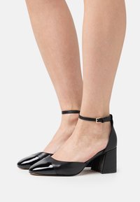 Zwarte gepatineerde leren blokhak schoenen met een spitse neus en een enkelbandje met een metalen gesp. Glad oppervlak en een elegant design.