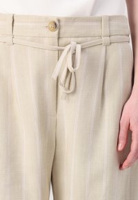 Pantaloni in lino beige chiaro con righe verticali bianche, dotati di chiusura con bottone e vita regolabile con coulisse. Tessuto strutturato.