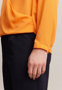 Bright orange blouse with long sleeves and a relaxed fit, paired with dark navy trousers. Features smooth fabric and a buttoned cuff detail.