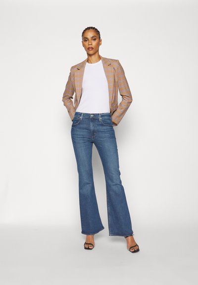 Blazer scozzese marrone e giallo su misura, t-shirt bianca, jeans a zampa di elefante blu e tacchi neri con cinturini, in piedi davanti a uno sfondo bianco.