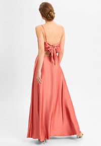 Luftiges korallenfarbenes Satin-Kleid mit dünnen Trägern und einer gebundenen Rückenpartie, das einen offenen Rücken und einen bodenlangen Rock hat. Glatte Textur.