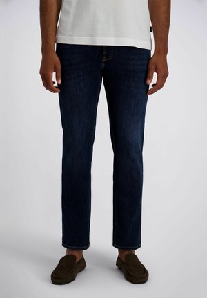 Mann trägt dunkelblaue Jeans mit geradem Bein, weißes kurzärmeliges Hemd und braune Slipper, steht mit entspannten Armen an den Seiten.