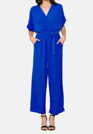 Kvinde iført en lys blå bredbent jumpsuit med korte ærmer, V-hals, taljebånd, lommer og sorte højhælede sko med ankelrem.