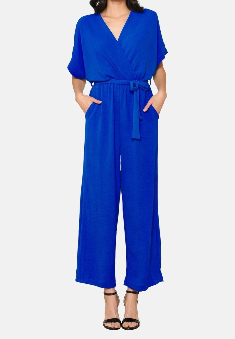 Femme portant une combinaison bleu vif à jambes larges avec manches courtes, col en V, ceinture à la taille, poches et talons hauts noirs à bride cheville.