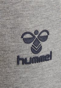 Šedá tkanina s texturovaným povrchem, která má tmavě námořnickou vyšitou logo a design včetně emblému připomínajícího včelu nad textem "hummel".