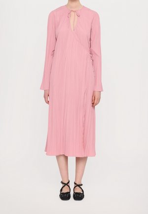 Rosa langärmliges Kleid mit Schlitz am Ausschnitt, plissierter Textur, asymmetrischem Design und seitlichen Bindebändern. Schwarze Schuhe mit überkreuzten Riemen.