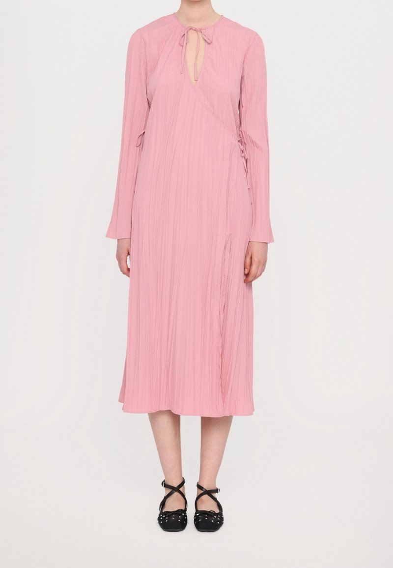 Robe rose à manches longues avec un col en trou de serrure, texture plissée, design asymétrique et liens latéraux. Chaussures noires à brides croisées.