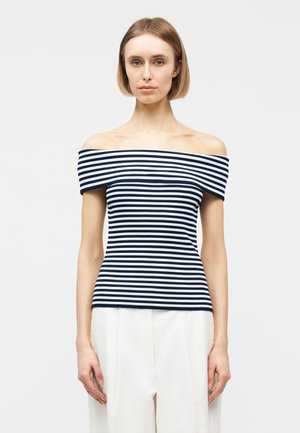 Frau mit kurzen blonden Haaren, die ein schulterfreies, marineblau-weiß gestreiftes Oberteil und lockere weiße Hosen trägt, vor einem schlichten Hintergrund stehend.