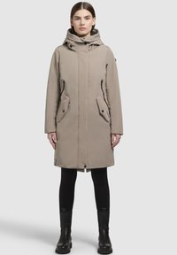 Beige waterdichte parka met hoge kraag, verstelbare capuchon, zijzakken en zwarte accenten. Gemaakt over zwarte leggings en laarzen.