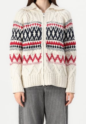 Pull blanc en maille avec fermeture éclair et motifs géométriques rouges et noirs, porté avec un pantalon gris rayé.