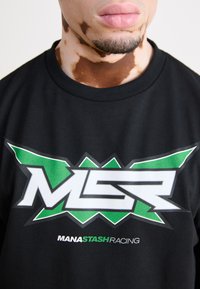 Schwarzes Baumwoll-T-Shirt mit einem großen weißen und grünen Grafikdesign, das "MSR" in fettem Text zeigt, und "MANASTASH RACING" darunter in kleinerer Schrift.