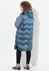Dicker, türkiser Weste mit Kapuze, die ein Chevron-Muster aufweist. Getragen über einem blauen Pullover, kombiniert mit dunklen Jeans und metallischen Schuhen.