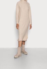 Robe-pull beige en maille avec un col roulé et des poignets côtelés, présentant des fentes latérales. Associée à des bottines taupe à semelle épaisse.