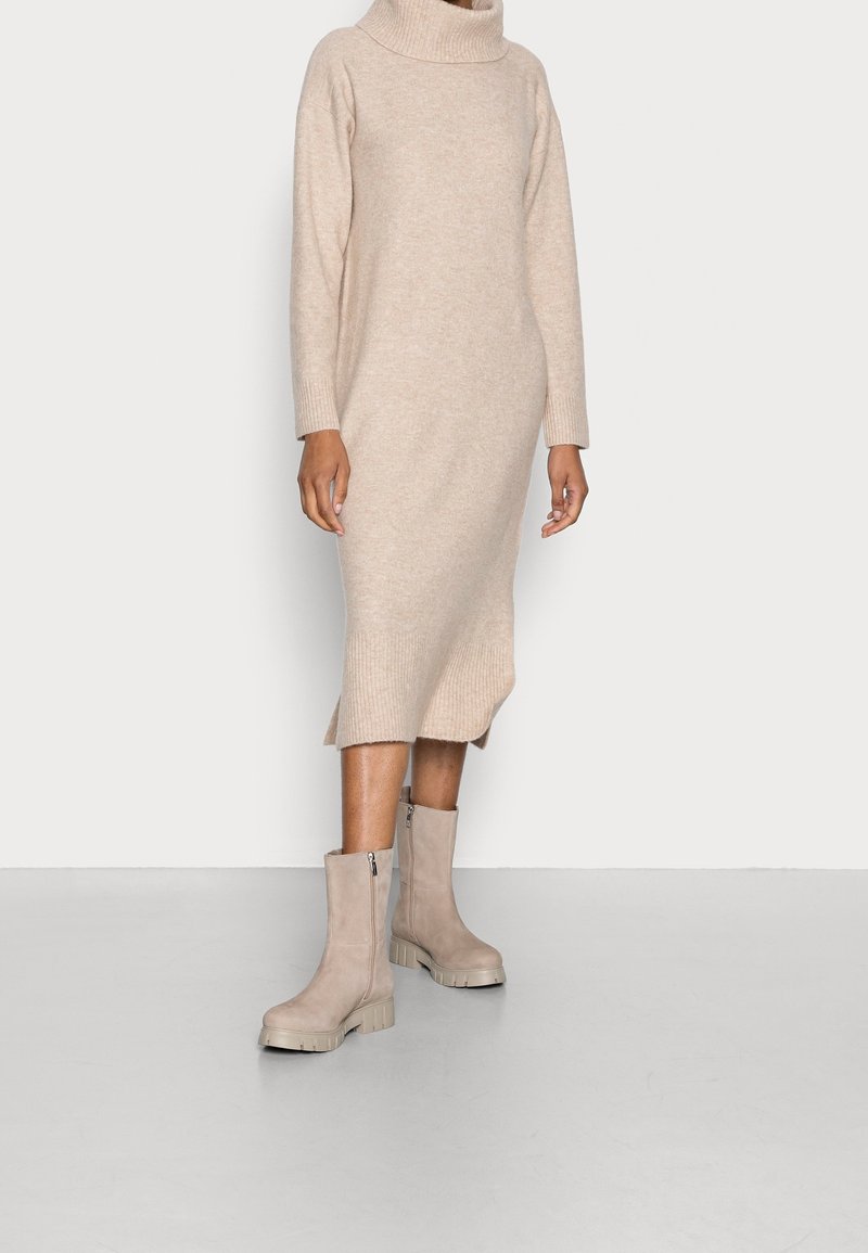 Robe-pull beige en maille avec un col roulé et des poignets côtelés, présentant des fentes latérales. Associée à des bottines taupe à semelle épaisse.
