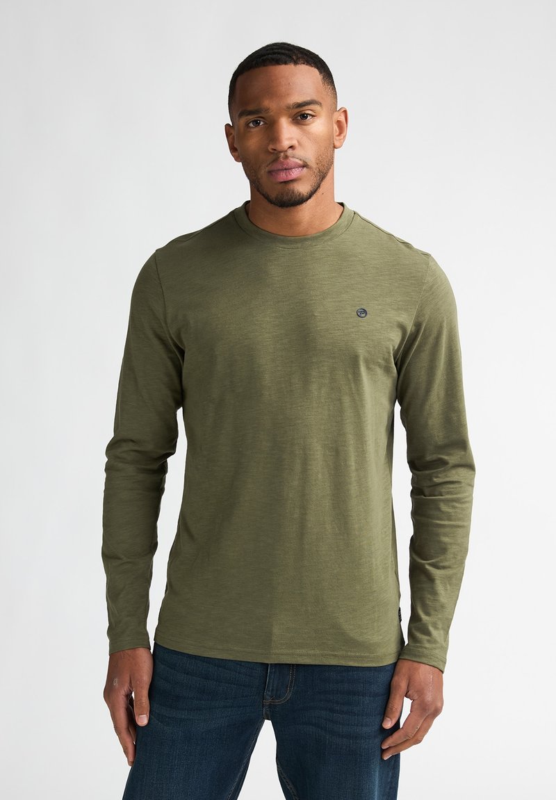 Zelené tričko s dlouhým rukávem a kulatým výstřihem s malým logem. Vyrobené z měkké, texturované látky. Kombinováno s modrými džínami.