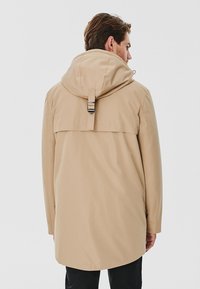 Beżowy wodoodporny płaszcz z kapturem, regulowanym paskiem i długimi rękawami, wykonany z gładkiego, lekkiego materiału, o prostym kroju.