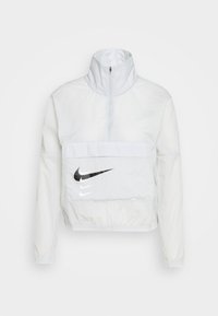 Veste Nike blanche avec col montant, fermeture demi-zippée et une poche avant ornée de logos noirs et blancs. Conçue en matériau léger.