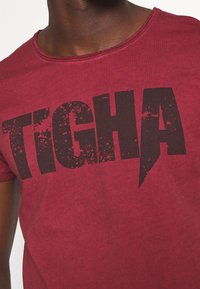 Camiseta corta roja de algodón con mangas cortas, que presenta un gran texto gráfico negro expresivo con un efecto desgastado.