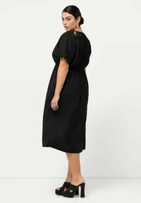 Vestido midi negro con detalles de encaje en las mangas, cintura elástica y corte holgado. La modelo lleva zapatos de plataforma negros. Fondo liso.