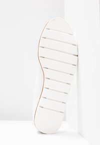 Baskets blanches avec une semelle en caoutchouc texturé et des rainures verticales. Présente un léger accent beige sur le bord et un logo en relief sur le côté.