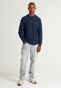 Sweatshirt blu navy con collo a barca e polsini a costine, abbinato a jeans azzurri chiari e sneakers beige. Design minimalista con accento del logo.