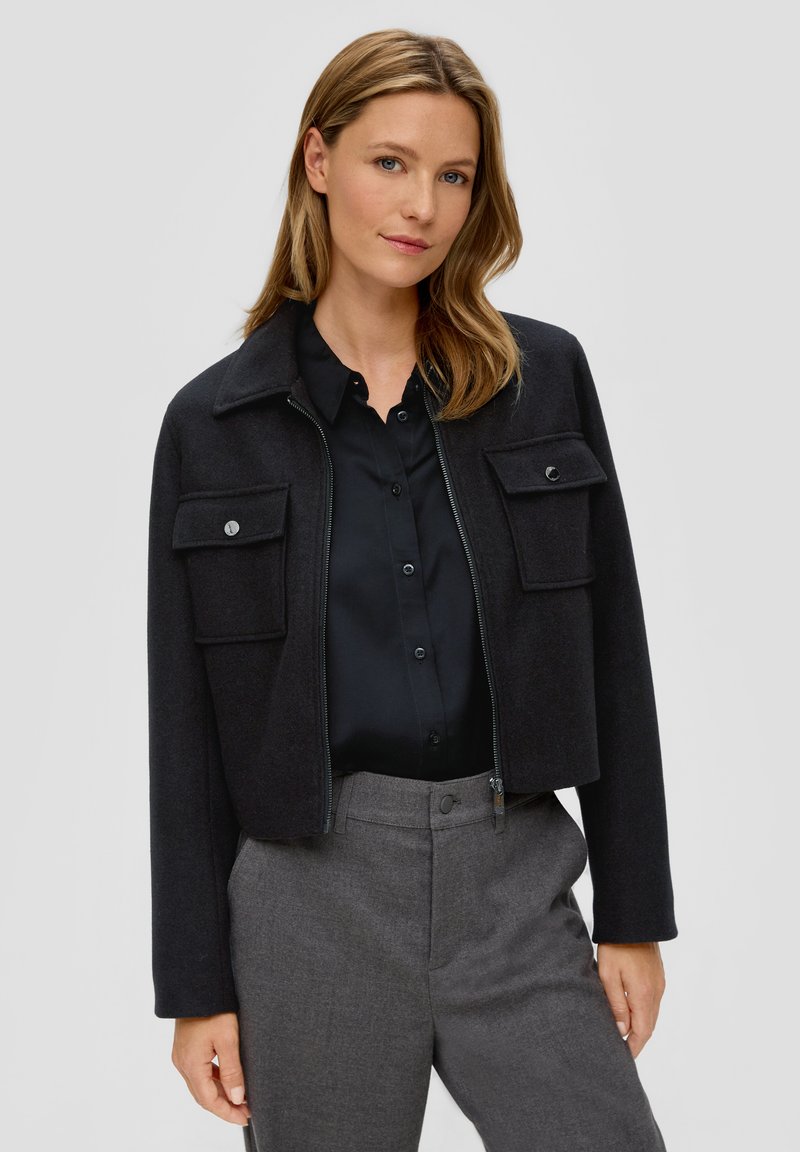 Czarna krótka kurtka z kołnierzem, dwiema kieszeniami na piersi i zamkiem błyskawicznym. Noszona na czarnej koszuli i szarych spodniach. Gładka faktura.