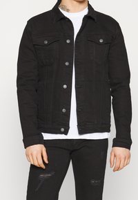 Svart denimjacka med krage, framknappar, bröstfickor och urtvättad detalj. Bärs över en vit t-shirt och svarta jeans.