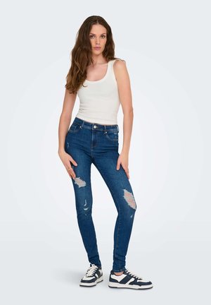 Jonge vrouw met lang bruin haar, gekleed in een wit mouwloos shirt, gescheurde blauwe strakke jeans en zwart-witte sneakers, staand.