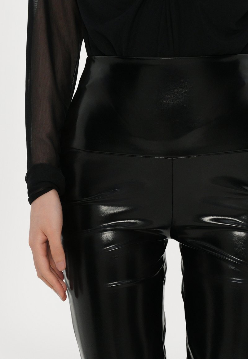 Leggings neri a vita alta realizzati in materiale lucido e elasticizzato. Presentano una texture liscia con dettagli leggermente stropicciati lungo le gambe.