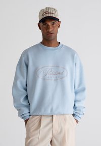 Sudadera azul claro con escote redondeado y hombros caídos. Presenta un logotipo bordado en rosa que dice "Prohibido" y estrellas. Combinada con pantalones beiges.