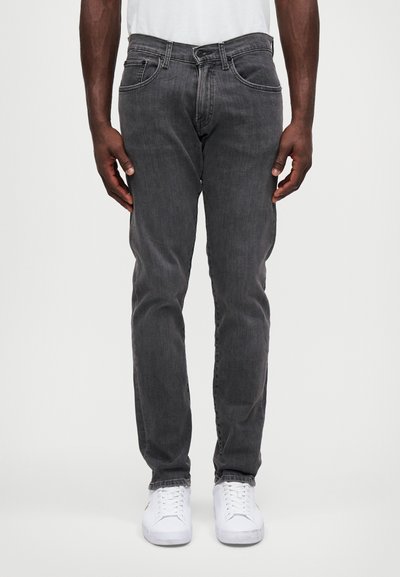 Vaqueros de mezclilla gris con ajuste slim, cinco bolsillos y un ligero efecto de desgaste. Combinados con zapatillas blancas, de pie contra un fondo sencillo.