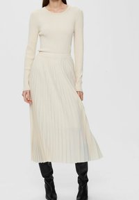 Top a maniche lunghe crema abbinato a una gonna midi plissettata in beige chiaro. Tessuto texturizzato, vita aderente e design fluttuante. Indossato con stivali neri fino al ginocchio.