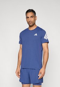 Camiseta deportiva azul de manga corta, con un pequeño logo blanco en el pecho y tres franjas blancas en cada hombro.