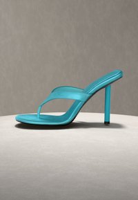 Τιρκουάζ σανδάλι με τακούνι σε σχήμα στρινγκ και λεπτό στιλέτο, τοποθετημένο σε κυκλική μπεζ επιφάνεια σε ουδέτερο φόντο.