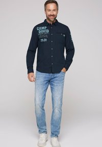 Camicia navy con bottoni, con testo e grafiche, taschino sul lato sinistro del petto, abbinata a jeans azzurri e sneakers bianche, in una vista a figura intera.