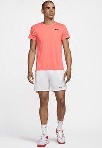 Nike sportinė apranga, kurią sudaro ryškiai koralų spalvos trumpomis rankovėmis marškiniai, balti šortai, balti kojinai ir sportbačiai su raudonais akcentais.
