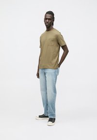 Tricou verde măsliniu cu buzunar la piept și logo mic, asortat cu blugi albastru deschis și pantofi sport negri cu tălpi albe.