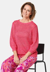 Rosa gestrickter Pullover mit losem, offenem Gewebe und Dreiviertelärmeln, kombiniert mit rosa Hosen im Blumenmuster. Sitzt auf einem schwarzen Stuhl.