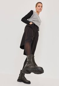 Gray knit vest over a black long-sleeve shirt, paired with black pleated shorts and green combat boots featuring laces and textured soles.