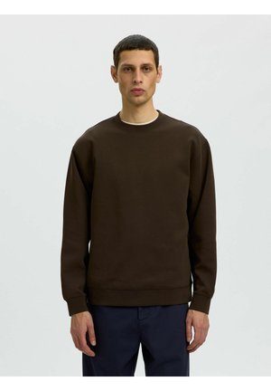 Man met kort haar die een effen donkerbruine trui en marineblauwe broek draagt, staat voor een effen lichte achtergrond.