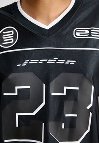 Svart idrettsjersey med hvit V-hals, stort tall "23" i hvitt, merkevaren logo, og strukturert stoff med horisontale linjer.