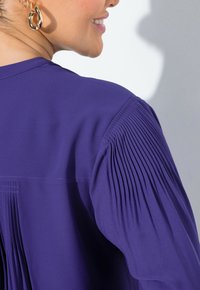 Blusa viola con dettagli plissettati sulle spalle, tessuto morbido con una superficie liscia e un design semplice del collo. Orecchini ad anello dorati visibili.