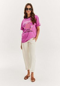 Růžové grafické tričko s tropickým designem, v kombinaci se krémovými kalhotami a hnědými sandály na řemínky. Jasný, uvolněný střih a krátké rukávy.
